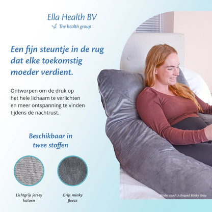 Een vrouw met een open boek zit op de bank, ondersteund door een Ella Sleeps zwangerschapskussen voor extra comfort.